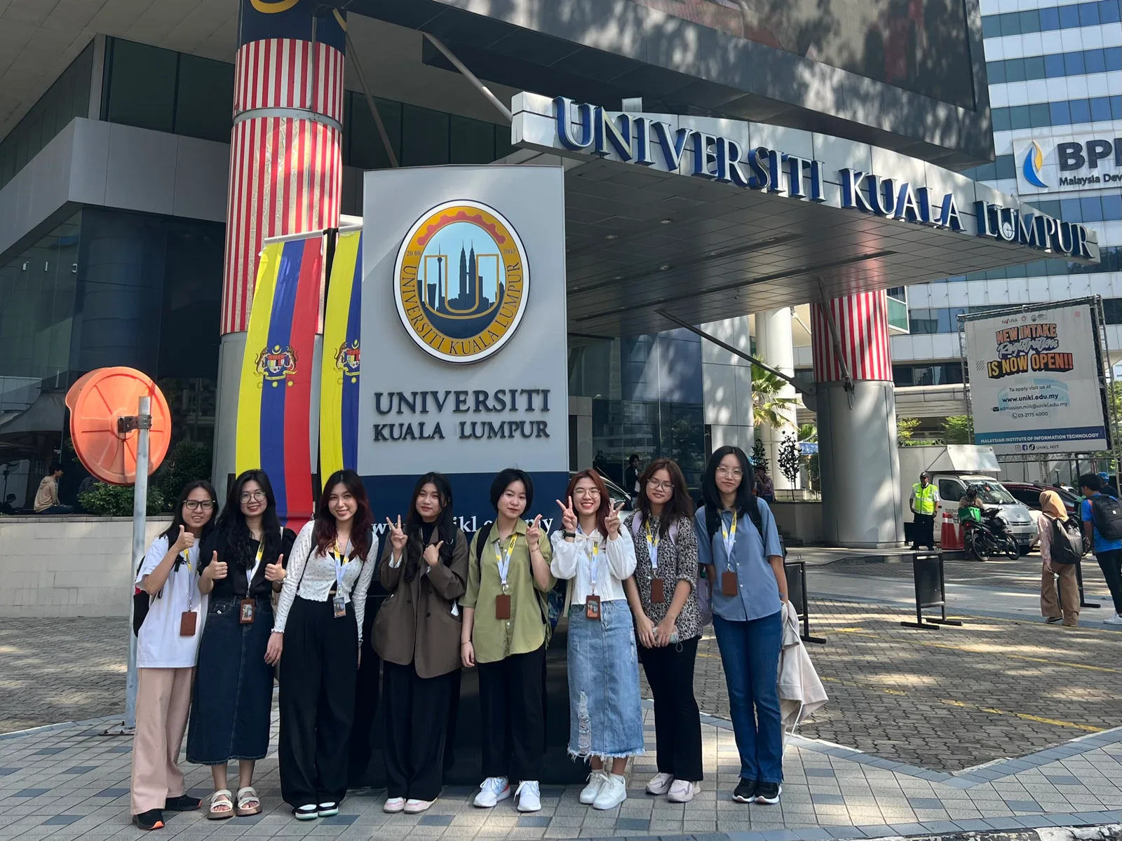 Nhóm bạn đứng trước biển tên trường và các lá cờ Malaysia